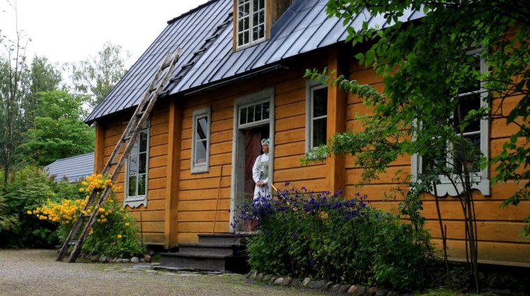 Keltainen vanha talo, kultapallot ja ukonhatut kukkivat, nainen vanhanaikaisessa asussa ovella.