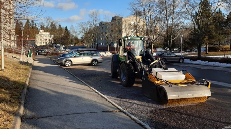 Traktori poistaa hiekoitushiekkaa paikoitusalueelta pyörvällä harjalla.