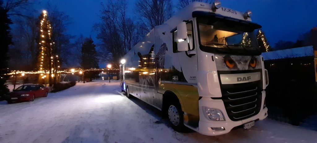 Kirjastoauto talvi-iltana, taustalla valaistu kuusi ja jouluvaloja RAntapusitossa.