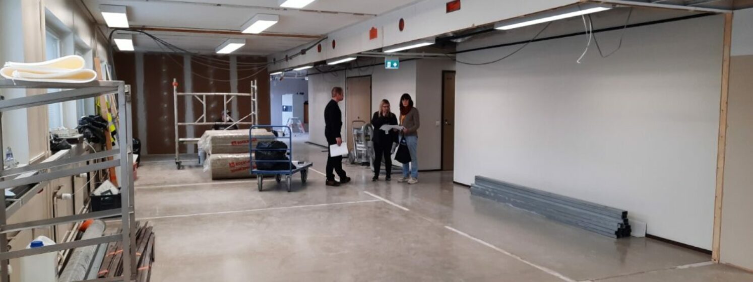 Rakenteilla oleva kirjaston väistötola, rakennustelineitä, kuljetuskärry ja henkilöitä paperit kädessä.