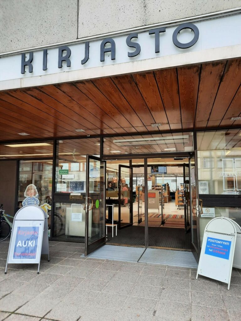 Kirjaston ulko-ovi auki, molemmin puolin ovea ständit, joissa toisessa teksti kirjasto on auki, toisessa ilmoitus poistomyynnistä.