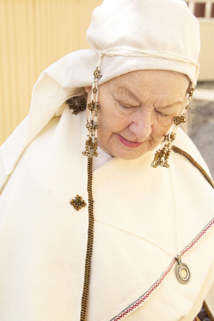 Kuva vanhemmasta rouvasta kansallis/muinaispukuun pukeutuneena. Kuvan keskiössä pukuun kuuluva päähine ja päähinen koristeelliset nauhat. Kuvat ottanut Silvi Kaarakainen