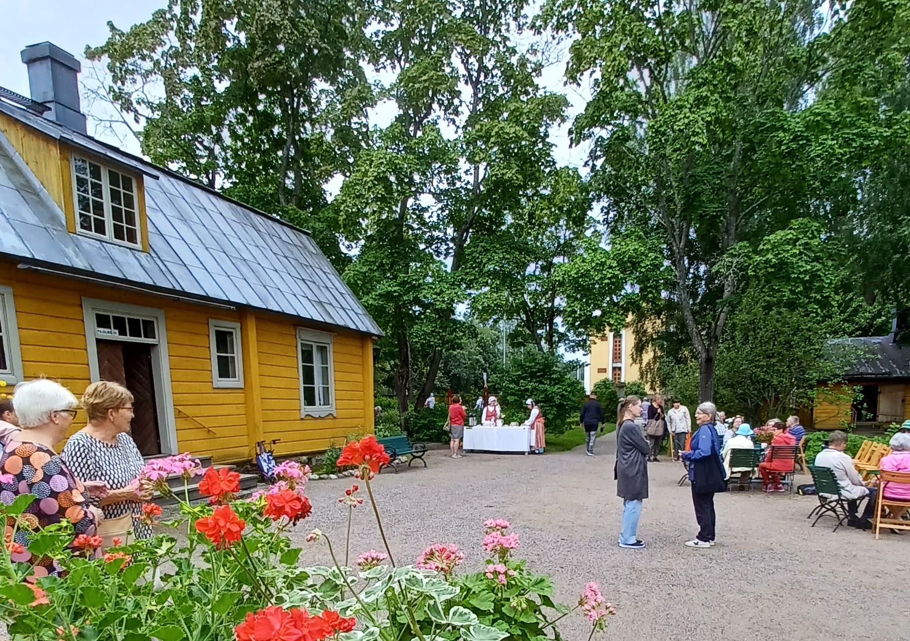 Vasemmalla keltainen vanha museorakennus, etualalla pelargoneja. Talon pihalla paljon ihmisiä.