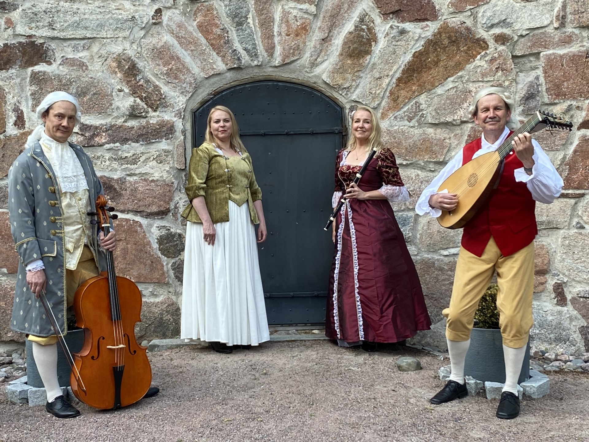 Neljä muusikkoa pukeutuneena 1700-luvun asuihin poseeraamassa kivimuurin edessä soittimien kanssa.