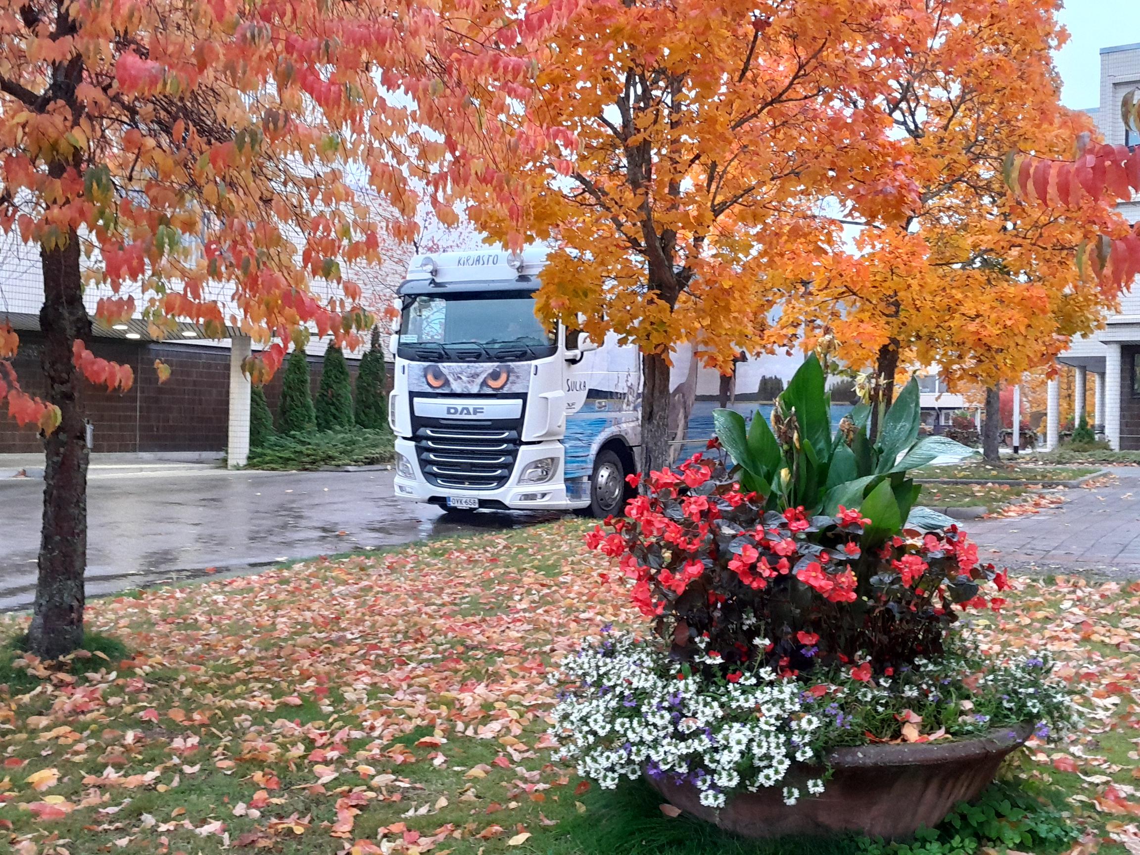Kirjastoauto edestä kuvattuna, ruskassa hekuvia puita ja värikäs kukkaistutus.