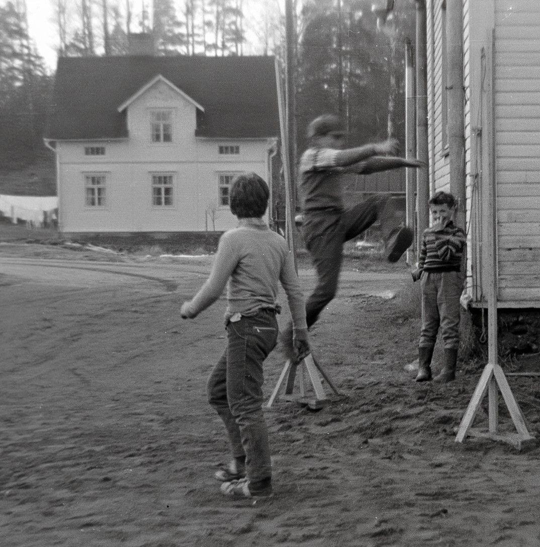 Mustavalkoinen kuva. Taustalla näkyy vaalea jyrkkäkattoinen puutalo. Etualalla on kolme poikaa hyppäämässä korkeutta.