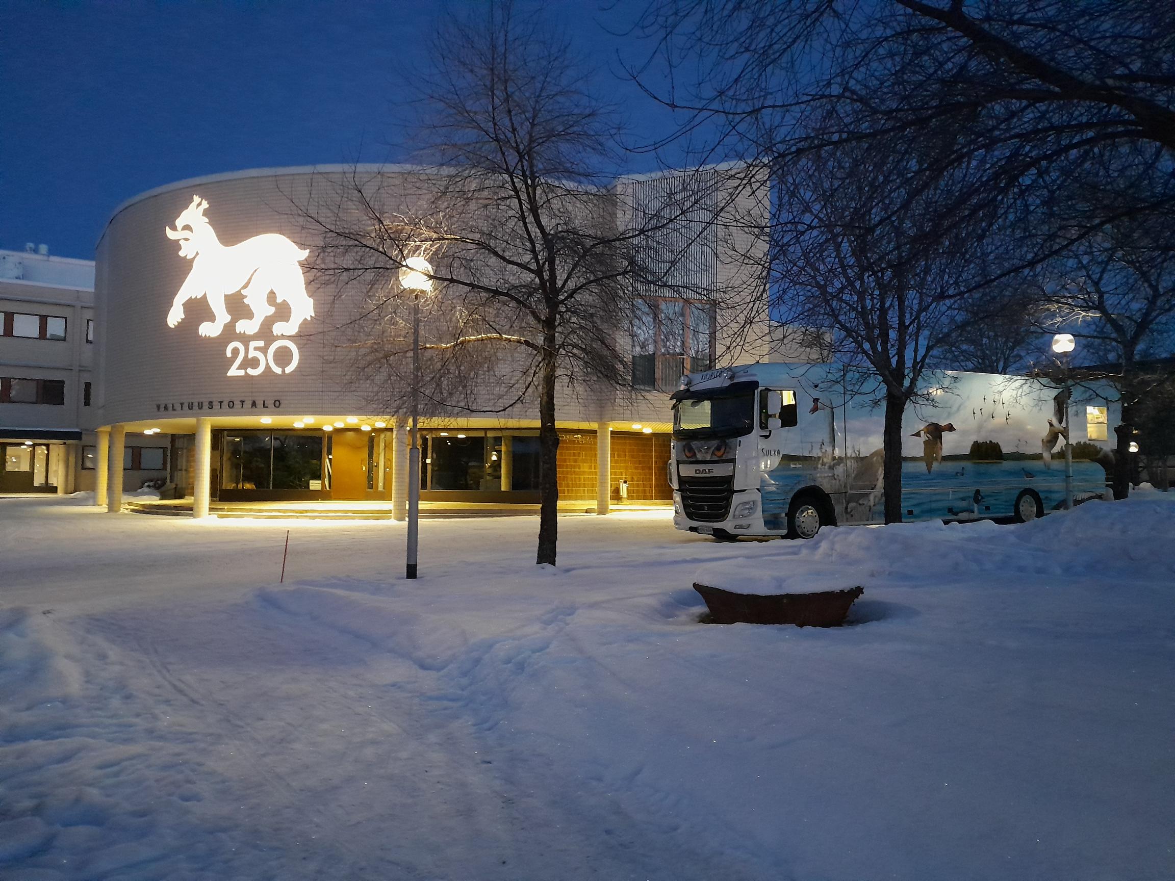 Lumisessa aamuhämärässä kirjastoauto pysäköity kaupungintalon eteen, kaupungintaloon heijastuu juhlavuoden logo ilves ja 250.