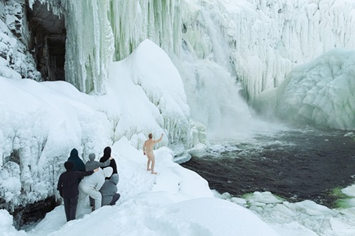 Jääputouksen reunalla alaston mies katsoo veteen osoittaen kädellään putousta. Tilannetta seuraa pieni ihmisjoukko vasemmassa reunassa.