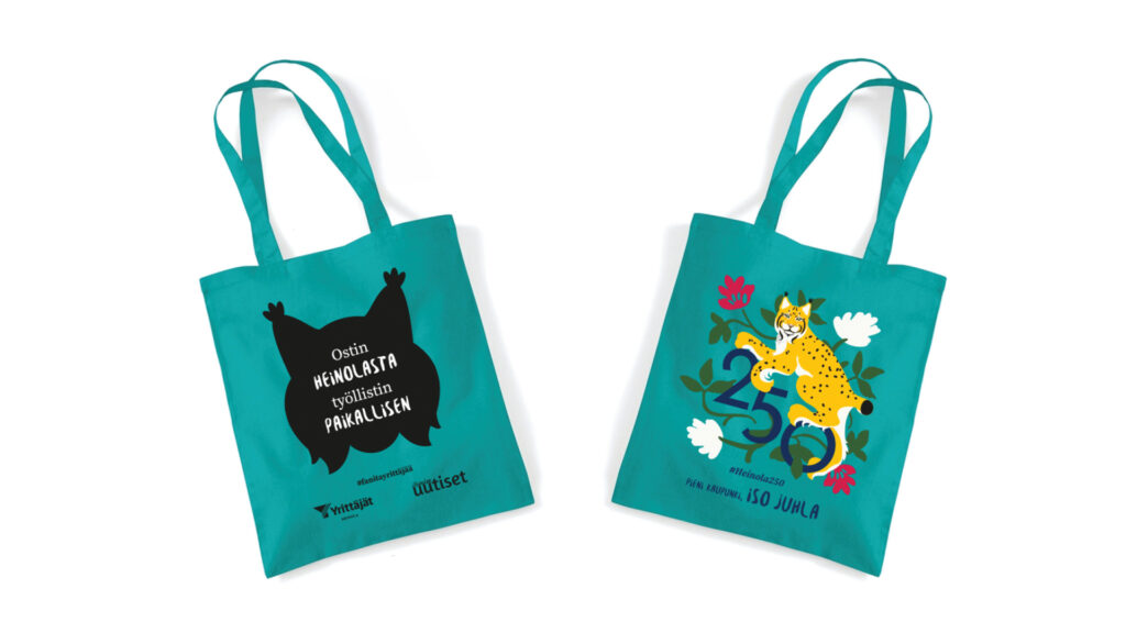 Kangaskassit joissa ilveksen pää ja slogan Osta Heinolasta, työölistä paikallinen. Toisella puolella juhlavuoden logo.