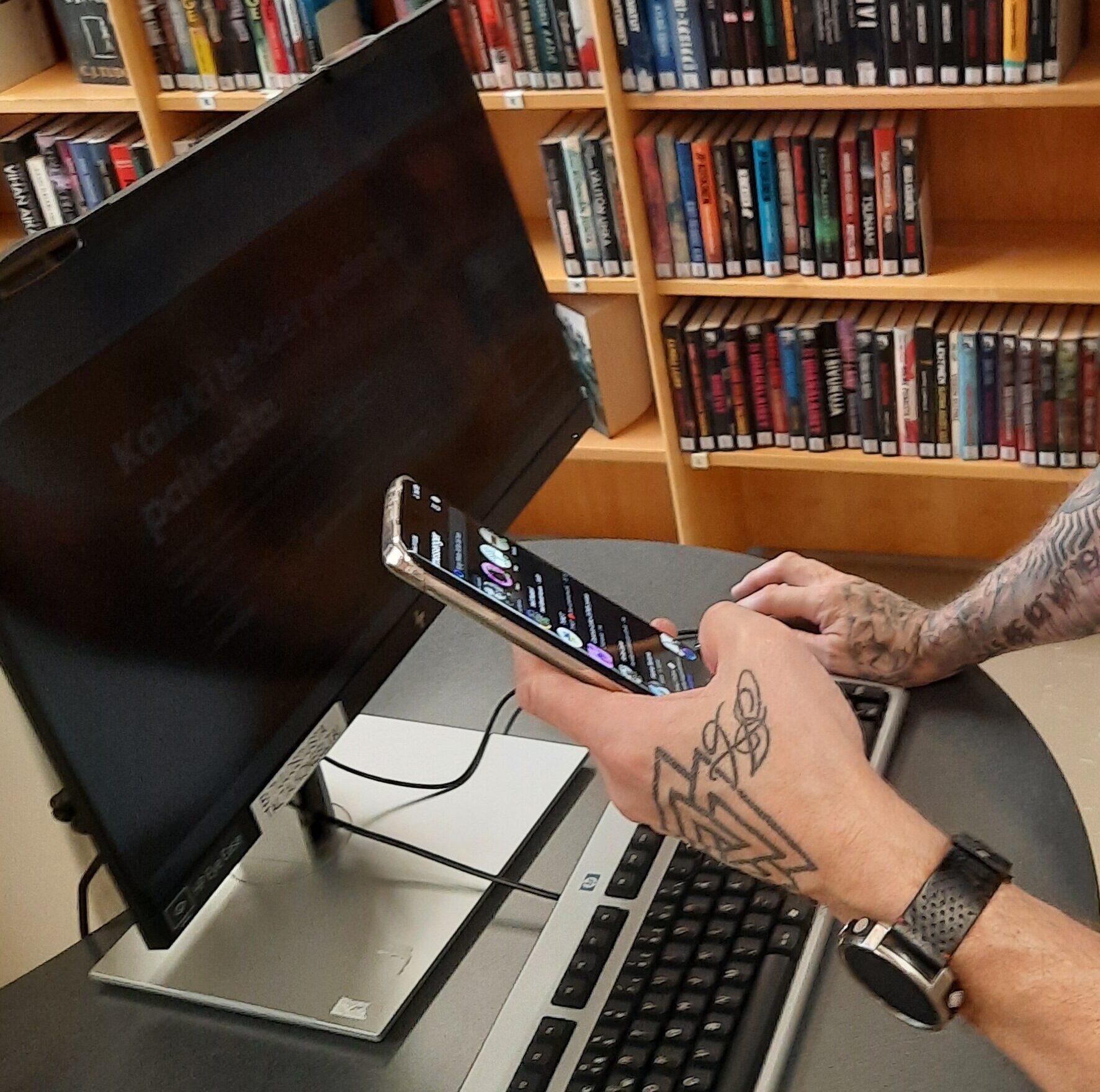 Tietoneneen näyttö, kirjoja hyllyssä ja älypuhelin tatuoidun henkilön kädessä.
