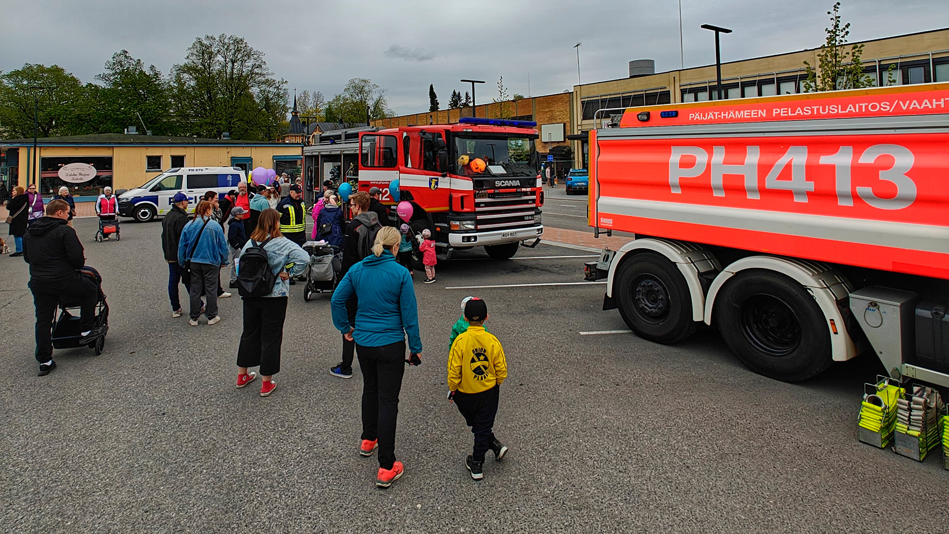 Ihmisiä, erityisesti lapsiperheitä on kokoontunut ulkoalueelle katsomaan paloautoja ja poliisiautoa, jotka on pysäköity torialueen pysäköintialueelle.