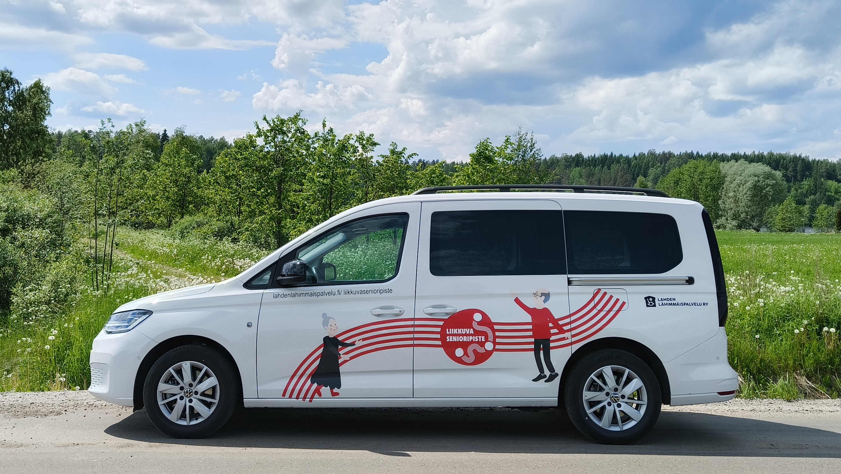 Kuvassa on Liikkuvan senioripisteen valkoinen tila-auto kesäisessä peltomaisemassa.