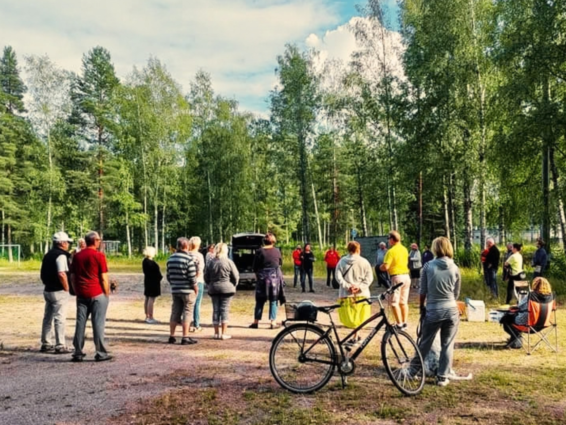 Ihmisiä seisomassa hiekkakentällä, taustalla puita.